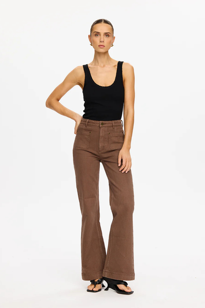 A full-length front studio photograph of the model posing with her right hand resting on her hip. The composition frames the complete outfit layout, showcasing the sculpting high-rise waistline, the close-to-body denim fit, the utilitarian front patch pockets with metallic rivet accents, and the wide-leg flare hemline hitting just above the floor.