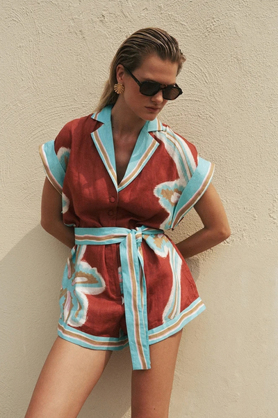 Outdoor lifestyle portrait of a model wearing sunglasses and the maroon and aqua printed playsuit against a textured wall.
