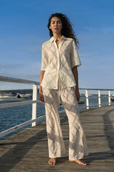 Full-length outdoor lifestyle shot of a model wearing the KIVARI Izzy Pant in Cream with the matching Izzy crochet shirt on a sunny wooden pier.