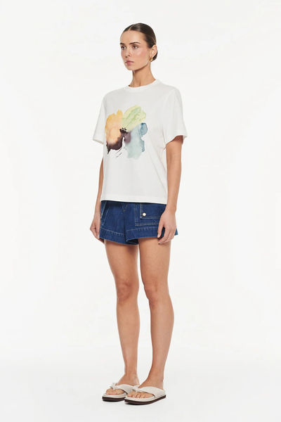 Studio three-quarter view of a model with her hair sleeked back, wearing a white organic cotton t-shirt with a colorful watercolor floral print, paired with dark blue denim shorts and beige slide sandals.