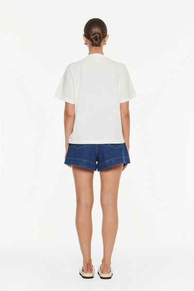 Studio rear full-length view of a model with a low bun, wearing a relaxed white short-sleeve organic cotton t-shirt and dark denim shorts.
