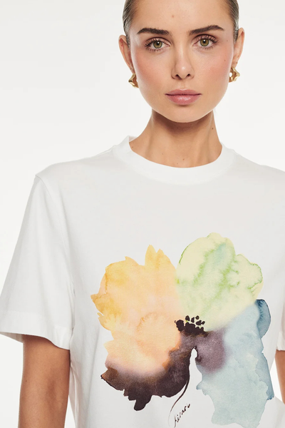 Close-up portrait of a model wearing gold earrings and an ivory crew-neck t-shirt featuring a large watercolor flower print in yellow, green, and blue tones.