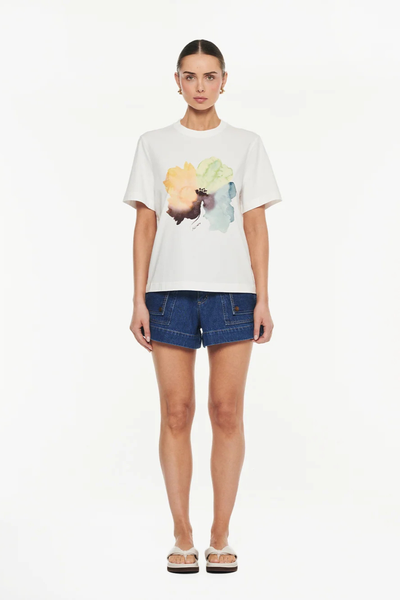 Full-length studio front view of a model wearing an ivory crew-neck t-shirt with a watercolor floral graphic, dark denim utility shorts, and cream slide sandals.