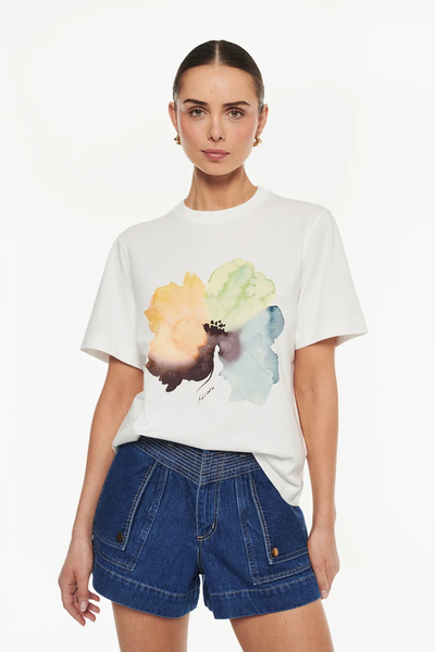 Mid-shot portrait view of a model wearing an ivory organic cotton t-shirt with a watercolor floral placement artwork, styled with dark blue denim shorts.
