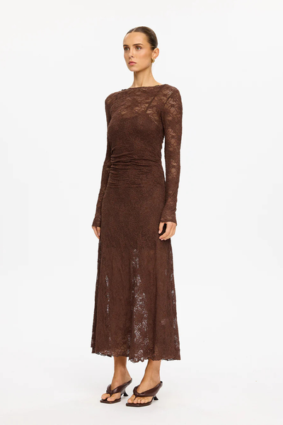 A full-length rear view of the model from behind against a plain white background. The image details the sheer quality of the chocolate lace fabrication below the inner jersey mini slip lining, the center-back seam structure, the continuation of the side waist ruching, and her hair styled in a neat high bun.