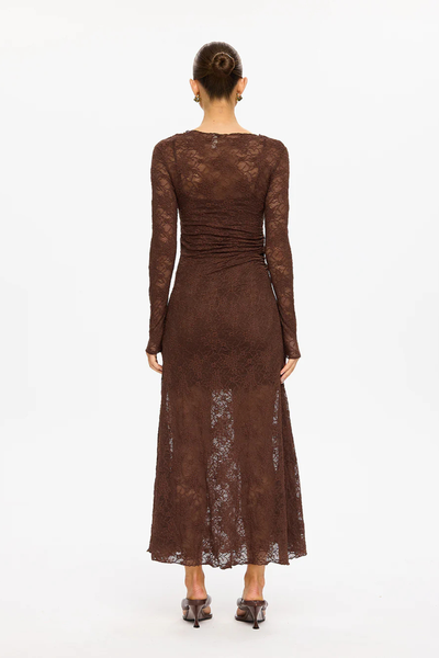 A full-length rear view of a studio model from behind, showcasing the clean design lines of the Marie Lace Maxi Dress in chocolate brown. The photograph illustrates the sheer stretch floral lace fabric extending down to a maxi length, contrasting with the opacity of the short, inner jersey mini slip lining. Key construction details visible include the fitted high neckline, slim full-length sleeves, a subtle horizontal gathering of ruching at the lower waist, and matching dark brown leather heeled sandals.