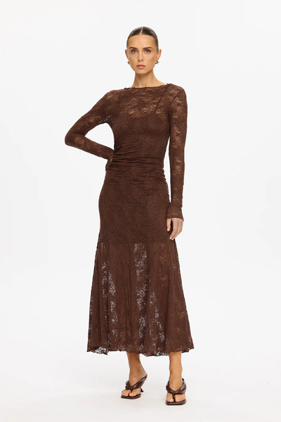 A full-length studio shot of the model posing with her right hand resting on her hip. The frame details the elongation of the slim-fit long sleeves, the fitted torso paneling, and the gentle flare of the semi-sheer maxi skirt hemline hitting just above the ankles.