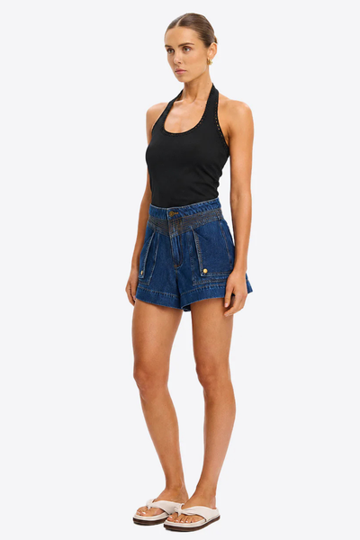 Three-quarter side angle view of a model styling the black ribbed Kivari Rhea Halter Top tucked into blue denim shorts and off-white slide sandals.