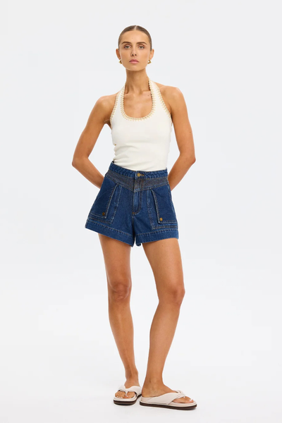 Full-length view of a model posing with hands behind her back in the cream-colored fitted Kivari Rhea Halter Top and dark denim shorts.