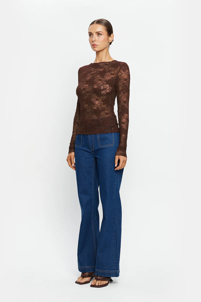 A full-length, three-quarter studio profile shot of a model standing at an angle against a clean white backdrop. She is wearing the semi-sheer chocolate brown stretch lace Victoria Top featuring full-length slim sleeves and a high neckline, paired with high-waisted wide-leg blue denim jeans with prominent front patch pockets.