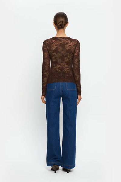 A full-length rear view of the studio model from behind against a white background, highlighting the back construction of the fitted chocolate brown lace top. The image showcases the sheer floral lace pattern across the back and shoulders, the clean seam lines, long slim sleeves, and how the hem sits flat over the waistband of the blue jeans.