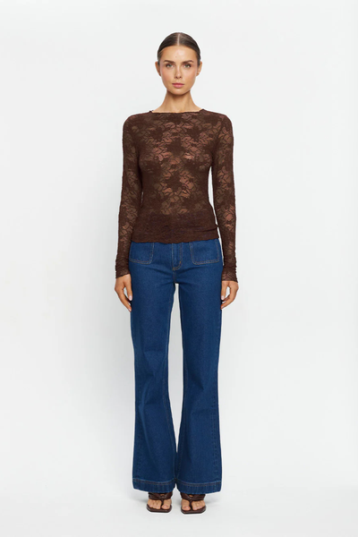 A full-length front studio photograph of the model standing straight against a plain white background. The composition captures the complete outfit styling, emphasizing the high-stretch fabrication, form-fitting silhouette through the torso, high neck shape, and full-length sleeves of the semi-sheer chocolate brown floral lace top tucked into wide-leg denim trousers.