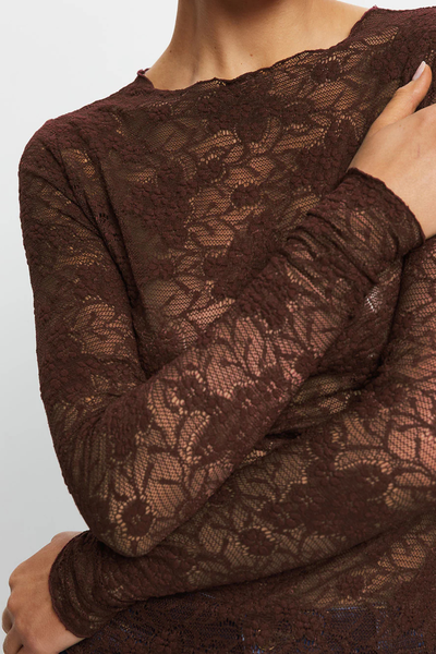 A detailed macro close-up shot focusing on the textile construction and upper right torso of the model. The image highlights the fine, intricate floral details of the 45% nylon, 50% cotton, and 10% elastane stretch lace, the delicate babylock edging along the high neckline, and the subtle texture of the gathered slim sleeves as her arms cross.