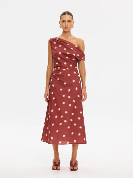 A full-length studio shot of a model standing straight-on against a white background, highlighting the asymmetric one-shoulder neckline and tailored silhouette of the Saro Midi Dress, paired with deep plum-colored kitten heel sandals.