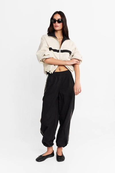 Full-length front view of a model standing with arms crossed softly, wearing the cream relaxed-fit jacket with black contrast details and black track pants.