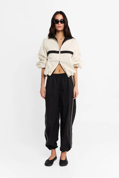 Full-length front view of a model wearing the cream and black spray jacket, black sunglasses, black track pants, and black flats, with the 2-way zipper partially open.