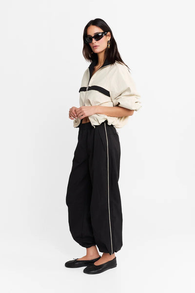 Three-quarter profile shot of a model wearing the cream and black spray jacket and black track pants, standing with hands clasped in front.