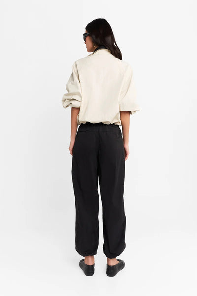 Full-length back view of a model wearing the cream Lennox spray jacket with a black contrast funnel collar, paired with black track pants and black flats against a plain white background.