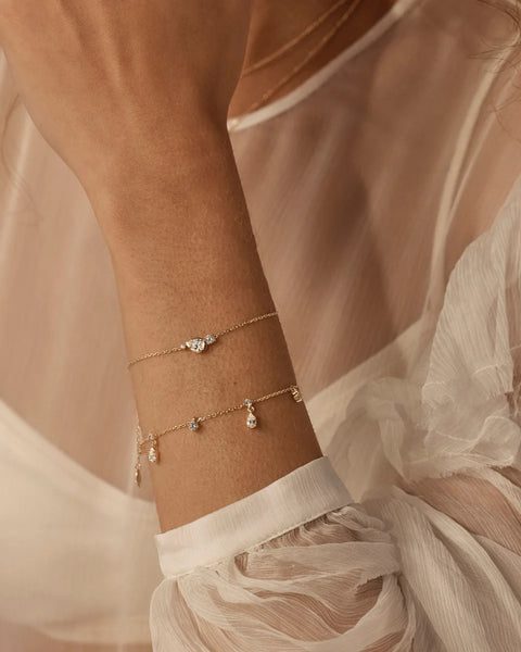 Model wearing the By Charlotte Quiet Radiance Bracelet in 18k gold vermeil stacked with a gold teardrop charm bracelet against a sheer white top.