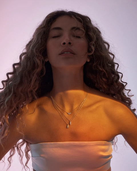 Campaign lifestyle shot of a model wearing layered 18k gold vermeil necklaces, including the By Charlotte Quiet Radiance Necklace, set under warm ambient lighting.