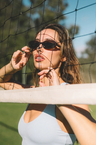 Outdoor campaign shot of a model wearing the Sito Shades Lourdes oval sunglasses in milky coco brown looking through a sports net on a sunny day.