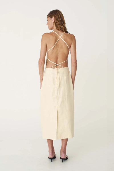 Studio rear view of a model wearing the butter creme linen midi dress, showcasing the apron-style silhouette, adjustable crisscross back straps, and centre-back hem vent.
