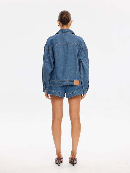 Full-length studio back view of a model showcasing the clean rear yoke line, dropped shoulder seams, and the tan leather logo patch stitched onto the hem of the oversized denim jacket.