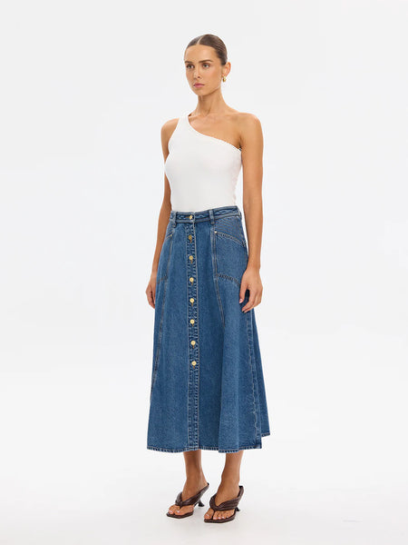 Full-length studio front view of a model posing with both hands in her pockets, highlighting the functional front pockets, detailed vertical paneling, and center button-down closure of the Kivari Rita Denim Midi Skirt.
