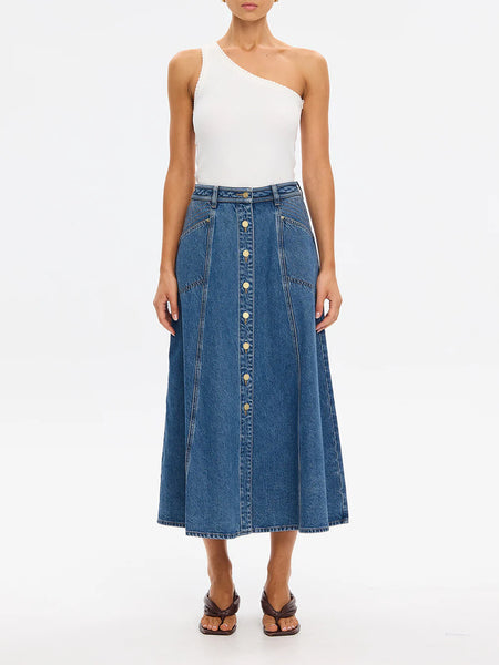 Full-length studio style campaign shot featuring a model posing from a three-quarter angle to show off the structured A-line silhouette of the Kivari Rita Denim Midi Skirt in Drift Blue, paired with an asymmetrical white one-shoulder tank top and brown strappy kitten heel sandals.