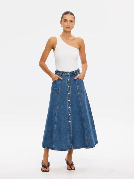A clean studio shot of a model standing straight-on against a white background, modeling the high-waisted vintage blue Rita Denim Midi Skirt styled elegantly with a tucked-in white one-shoulder top and dark brown heeled sandals.