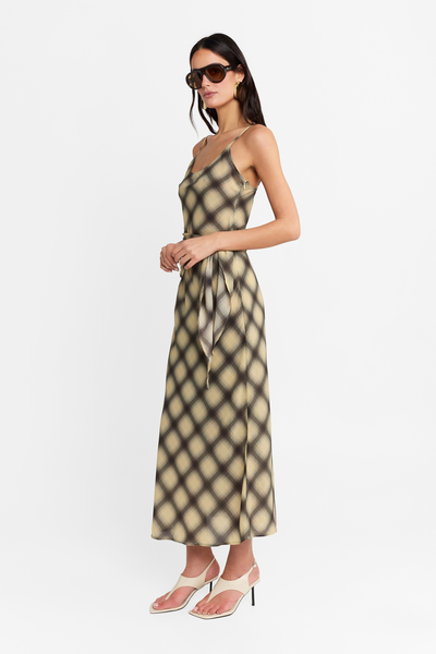 Full-length studio side profile view of a model wearing the check maxi dress featuring a scoop neckline, adjustable spaghetti straps, and a wrap-around waist tie, paired with cream-colored heeled sandals and sunglasses.