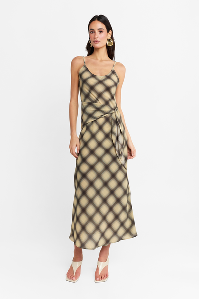 Full-length front studio view of a model wearing the check maxi dress with a scoop neckline, wrap tie, and midi-to-maxi length hem, styled with cream sandals and statement earrings.