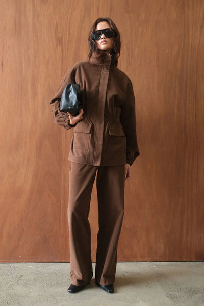 An editorial lifestyle portrait of a model posing against a warm wood-paneled wall. She is wearing the oversized espresso Amari utility jacket zipped up, paired with a small black leather pouch bag, dark sunglasses, and matching trousers for a modern streetwear look.