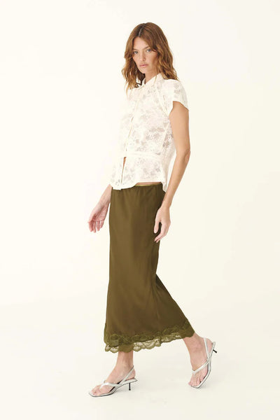 Full-length studio walking shot of a model wearing the ROWIE Marina Silk Midi Skirt in Olive with silver kitten heel sandals and an off-white lace top, highlighting the body-skimming silhouette and fine lace detailing.