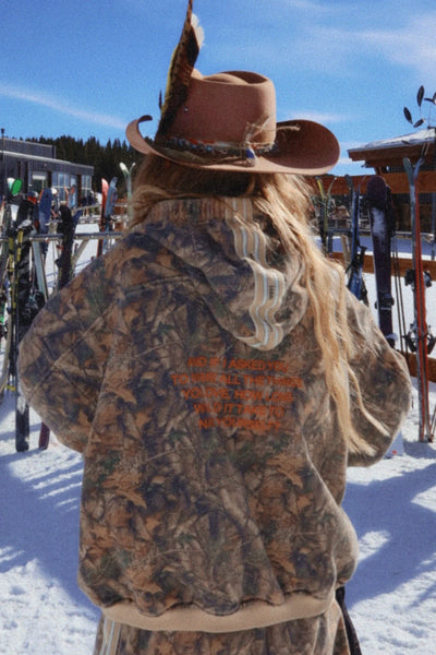 A full-length shot from the back on a model wearing the Name Yourself Hoodie, showcasing the large graphic statement artwork featuring a bold black and cream design inspired by adventure, along with the distinct contrast taping running down the back of the hood.