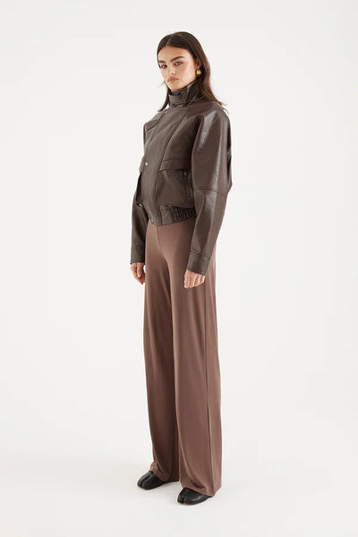 A full-length rear view of the model facing away from the camera against a plain white background. The shot highlights the utilitarian back design of the short jacket, showcasing the horizontal back flap panel stretching across the upper shoulders, the complex raglan sleeve paneling with distinct seam details, and the thick, gathered elasticated waistband finish.