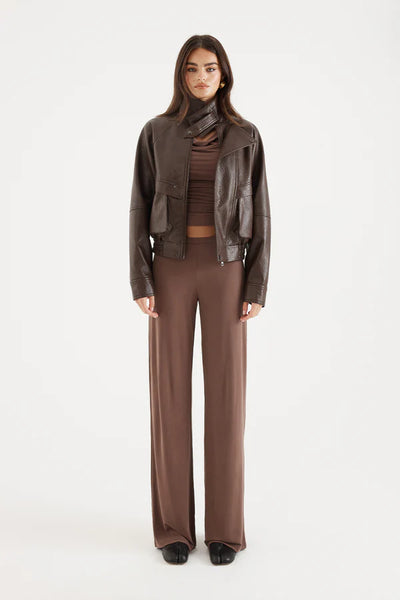 A full-length front studio photograph of a model standing at a slight three-quarter angle against a white backdrop. She is wearing the closed chocolate brown technical outerwear piece zipped up, showing how the boxy fit, asymmetric front panel, and elasticated hem interact with fluid, high-waisted wide-leg brown knit trousers and black tabi-toe shoes.