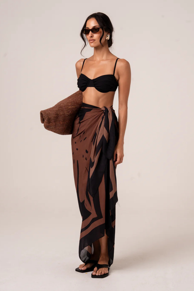 A full-length front studio view of a model wearing the Ochre Lane Alba Sarong in the Fiore print, tied at the hip to showcase the fluid, semi-sheer drape over swimwear and the elegant side-slit effect.
