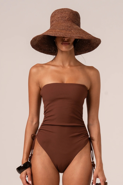 Front view of a model wearing the cocoa raffia bucket hat and a brown strapless one-piece swimsuit with side-tie ruched details.