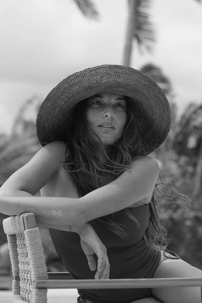 Black-and-white lifestyle shot of a model relaxing in a chair outdoors while wearing the raffia bucket hat and swimsuit.