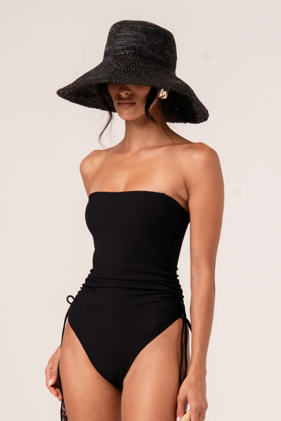 Front view of a model wearing the noir raffia bucket hat and a black strapless one-piece swimsuit with side ruching and ties.