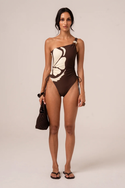 Full-length front view of a model wearing the asymmetric one-piece swimsuit in warm chocolate and cream ribbed fabric, featuring a beaded single-shoulder strap and side ties, accessorized with flip-flops and a small woven bag.