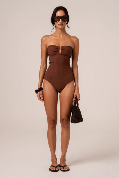 Full-length studio front view of a model wearing dark sunglasses, flat sandals, holding a dark woven mini bag, and wearing a chocolate brown ribbed strapless one-piece swimsuit.