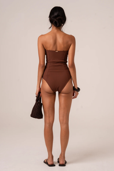 Studio rear full-length view of a model with her hair in a low bun, wearing flat slide sandals and a chocolate brown ribbed strapless one-piece swimsuit with full back coverage.