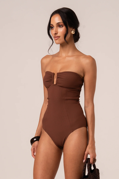 Studio three-quarter front shot of a model holding a dark woven bag, wearing gold statement earrings and a chocolate brown ribbed strapless bandeau one-piece swimsuit with vertical seams and a V-wire neckline.