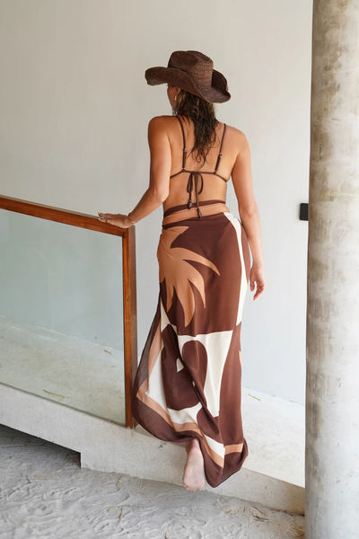 Rear view of a model walking up steps in a beachside villa wearing the semi-sheer Sombra Sarong as a maxi wrap skirt. The back layout reveals the large-scale abstract graphics in deep chocolate and warm cream, paired with a string triangle bikini top and a woven straw cowboy hat.