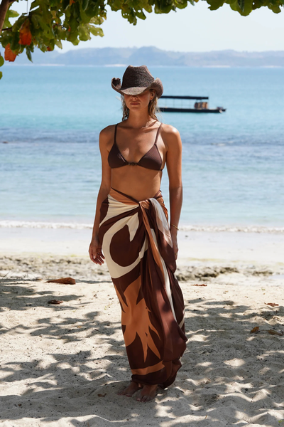 Front studio portrait of a model showcasing the multi-tonal Sombra Sarong tied as a high-waisted wrap maxi skirt. The semi-sheer, lightweight resort wear piece features an earthy palette of deep brown, tan, and cream abstract shapes, accessorized with a dark chunky bangle and black slide sandals.