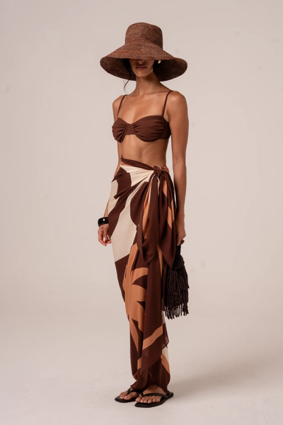 Full-length studio shot of a model wearing the Ochre Lane Sombra Sarong tied at the waist as a maxi skirt. The sarong features a bold, abstract graphic print in chocolate brown, warm cream, and tan, styled with a coordinating chocolate brown underwire bikini top and a wide-brimmed brown raffia sun hat.