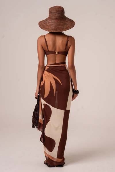 Full-length rear view of the Ochre Lane Sombra Sarong tied at the side hip, highlighting the fluid, semi-sheer drape across the back. The abstract floral-inspired graphic print shows large paneling in chocolate brown and cream, paired with a brown textured bikini top and a structured wide-brim sun hat.
