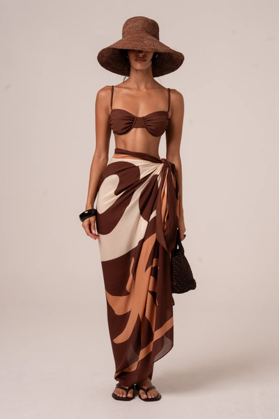 Front studio portrait of a model showcasing the multi-tonal Sombra Sarong tied as a high-waisted wrap maxi skirt. The semi-sheer, lightweight resort wear piece features an earthy palette of deep brown, tan, and cream abstract shapes, accessorized with a dark chunky bangle and black slide sandals.